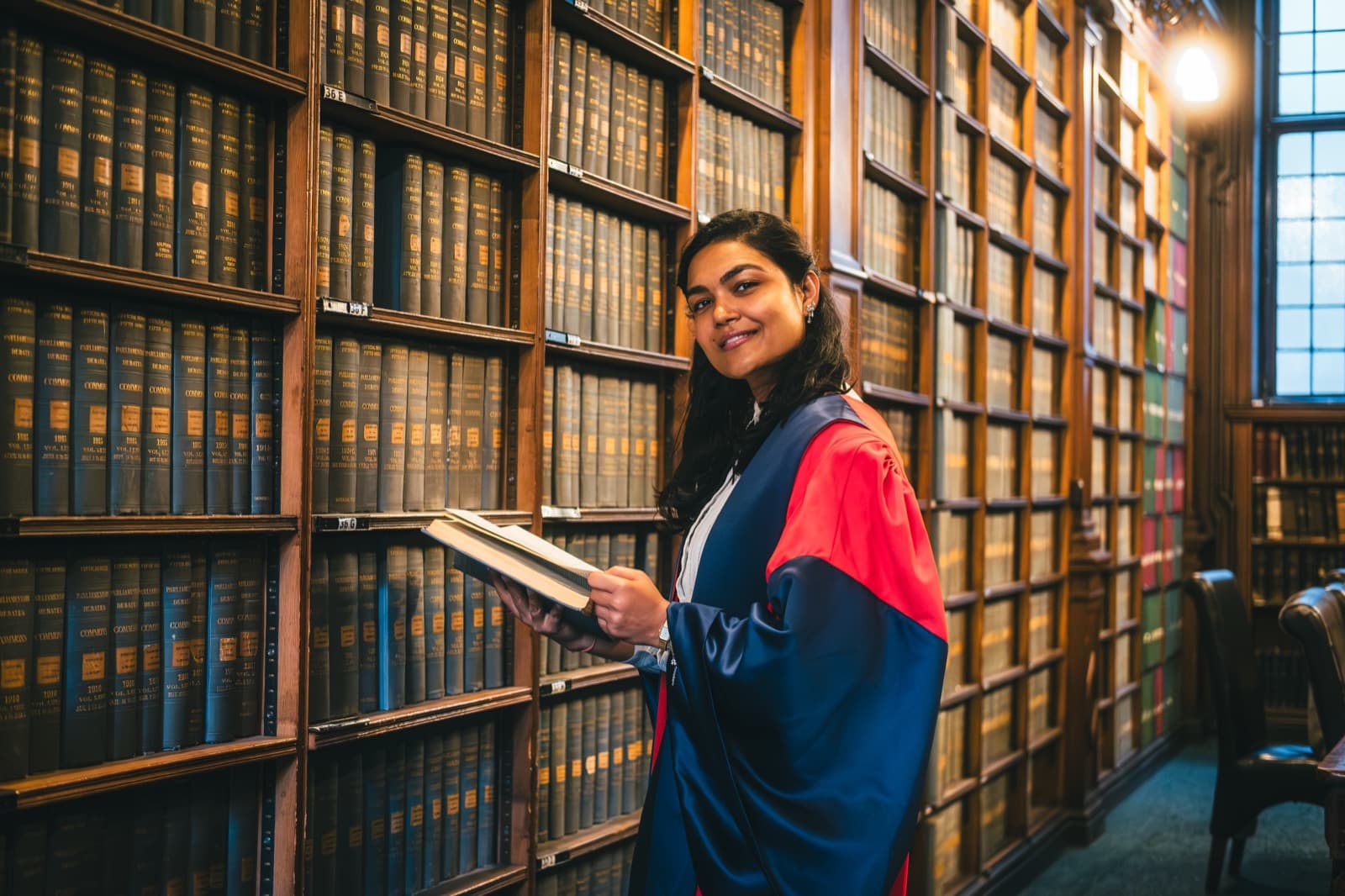 Dr Ayushi Agarwal in Oxford doctoral robes in a library of bound parliamentary volumes